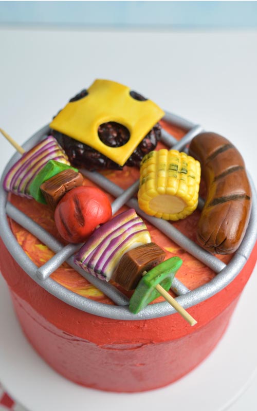Red buttercream barbecue grill cake with fondant sausage, corn, kebabs, burger with cheese.