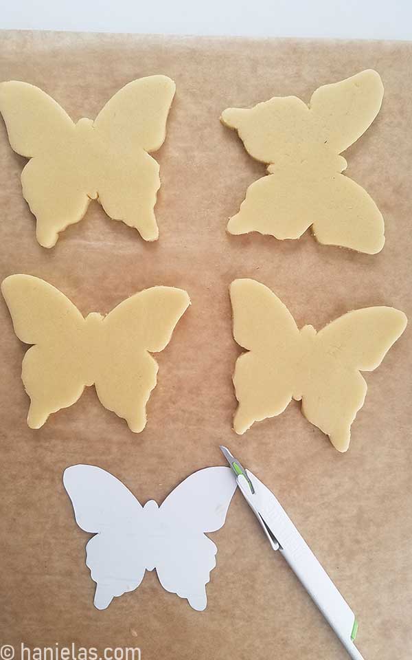 Unbaked butterfly cookies on a brown parchment along with a cookie cutter template and a pen blade knife.