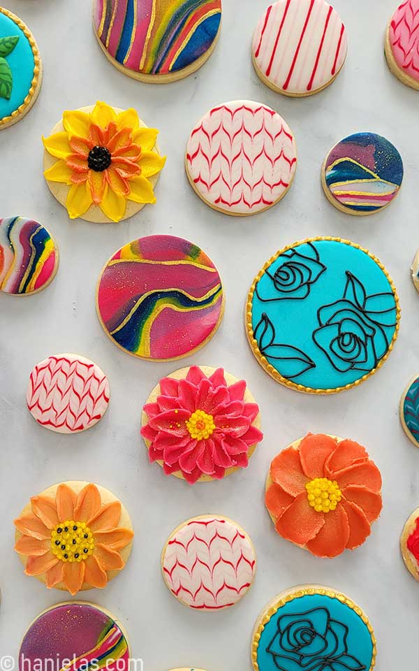 Round cookies decorated with fondant, royal icing, glaze, and buttercream displayed on marbled slab.