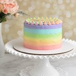 Rainbow frosted cake displayed on milk glass cake stand.