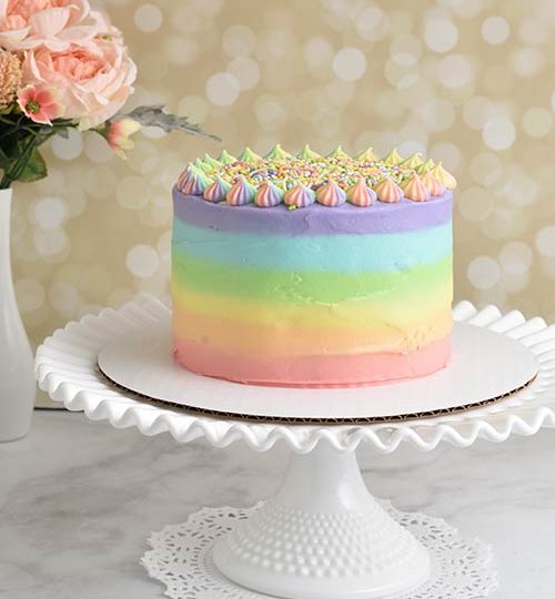 Rainbow frosted cake displayed on milk glass cake stand.