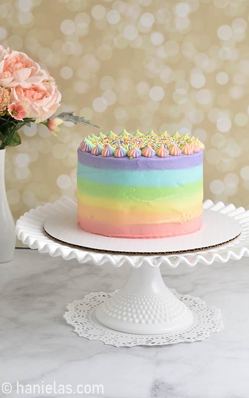 Rainbow frosted cake displayed on milk glass cake stand.