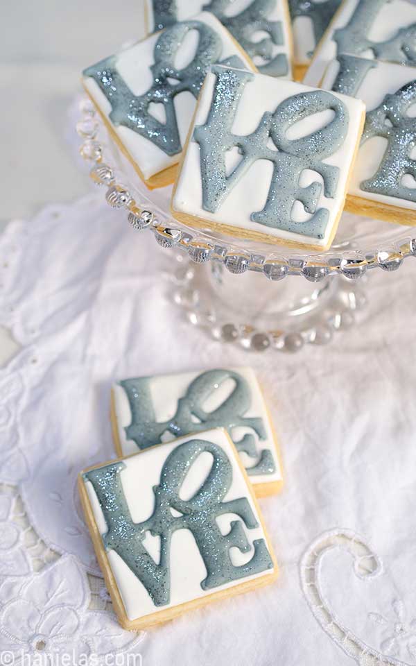 Square cookies decorated with a white icing base and topped with a gray royal icing transfer, stacked on a clear cake stand.