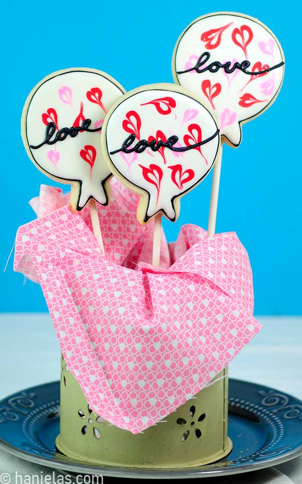 Decorated balloon cookies on sticks displayed in a small container.