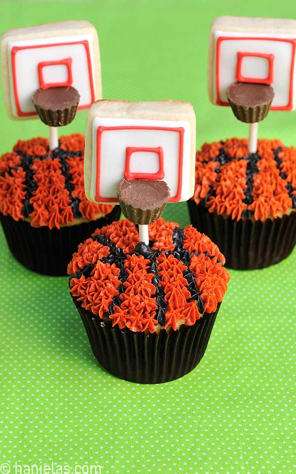 Cupcakes decorated with black and orange buttercream that look like basketballs with a cookie pop inserted in them, displayed on a green cloth.