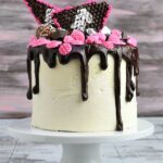 Cake decorated with thick chocolate drips, chocolate diamonds, and silver leaf on a white cake stand.