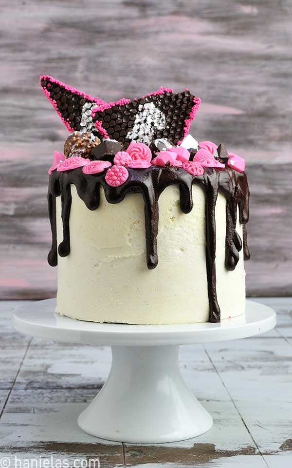 Cake decorated with thick chocolate drips, chocolate diamonds, and silver leaf on a white cake stand.