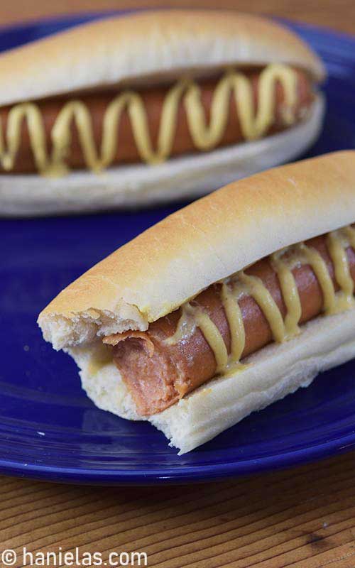 Baked bread bun filled with cooked hot dog and drizzled with mustard.