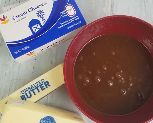 Thick caramel in bowl, block of cream cheese and stick of butter on a kitchen counter.