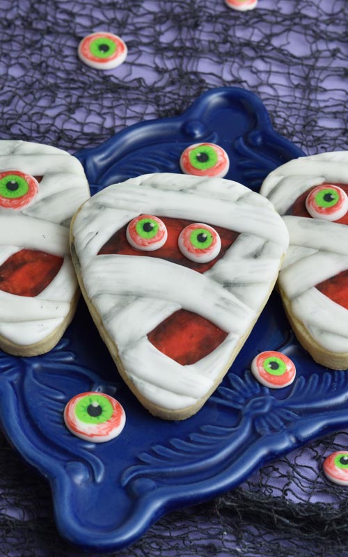 Decorated mummy cookies on a blue plate surrounded with monster royal icing eyes.