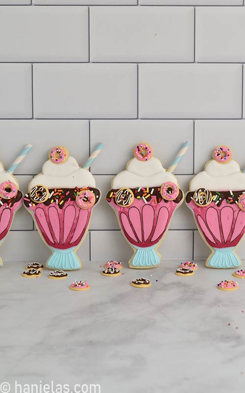 Cookies decorated with royal icing in a shape of milkshake displayed leaning towards the tile backsplash.