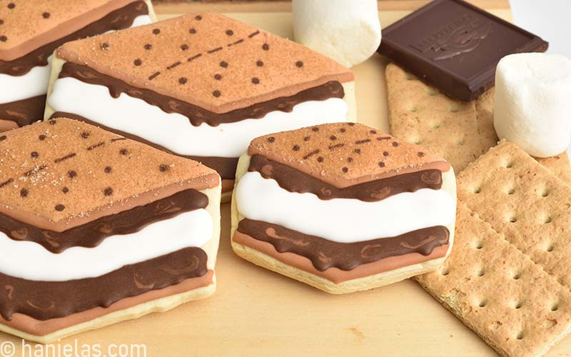 Royal icing decorated sugar cookies that look like s'mores, displayed on a wooden cutting board.