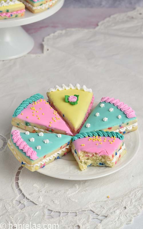 Decorated cookie that look like cake slices on a white plate with one cookie bitten.