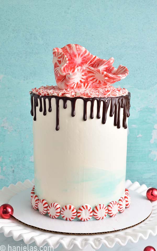 White cake with chocolate drips decorated with crushed peppermint candies on a white milk glass cake stand.