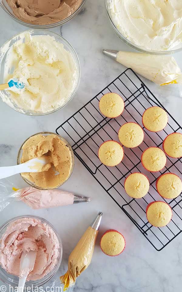 Undecorated cupcakes and buttercream in bowls on a marbled counter top.