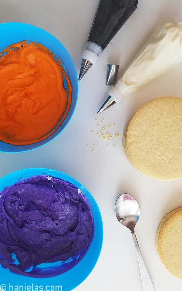 Orange and purple buttercream in bowls, piping bags filled with white and black buttercream on a kitchen counter.