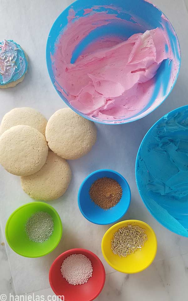 Pink and blue buttercream in bowls with undecorated cookies and sprinkles on a side.