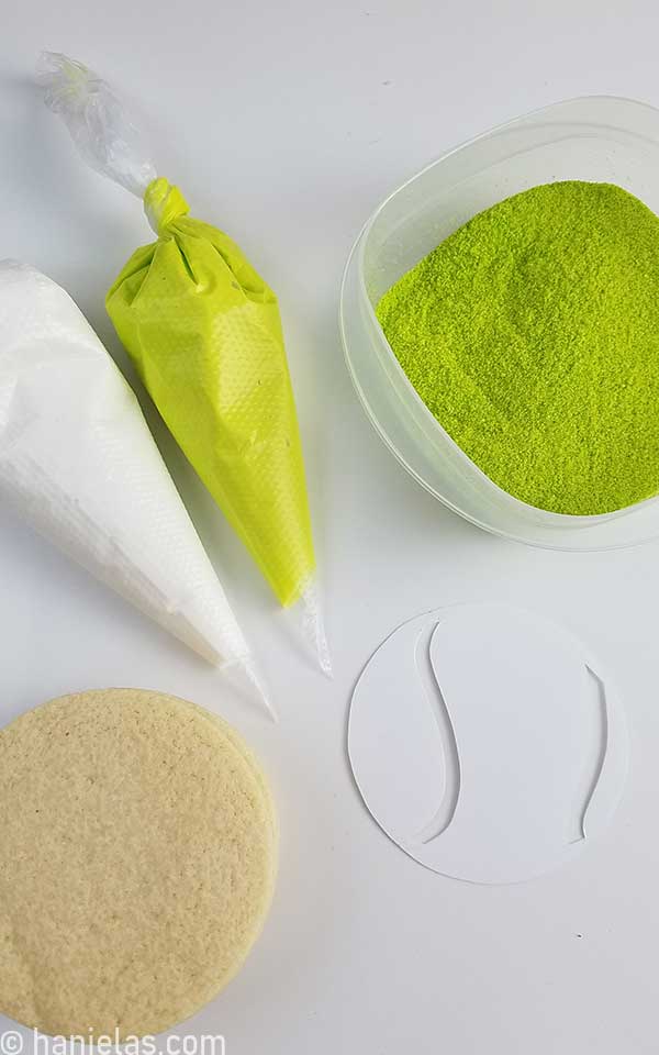 Two piping bags filled with icing, a small bowl with colored sugar and a round cookie on a counter.