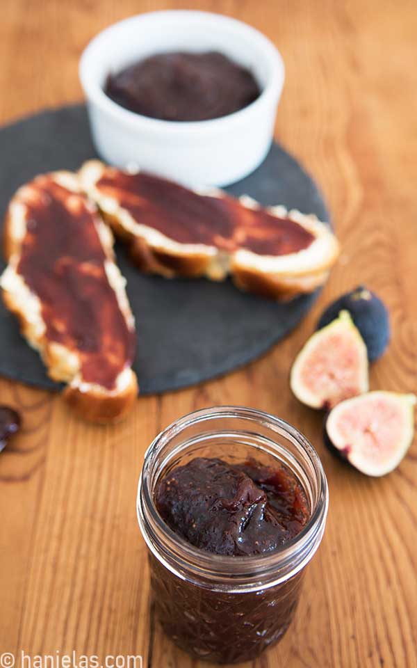 Wood board, black plate with bread with fig jam, fresh figs and a small jar with jam.