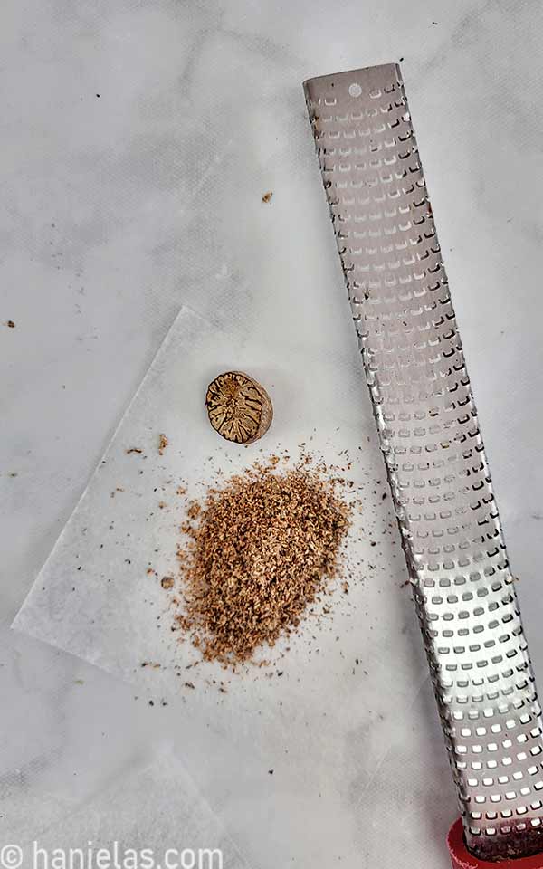 Half of the nutmeg, grated nutmeg and microplane zester on a marbled counter.