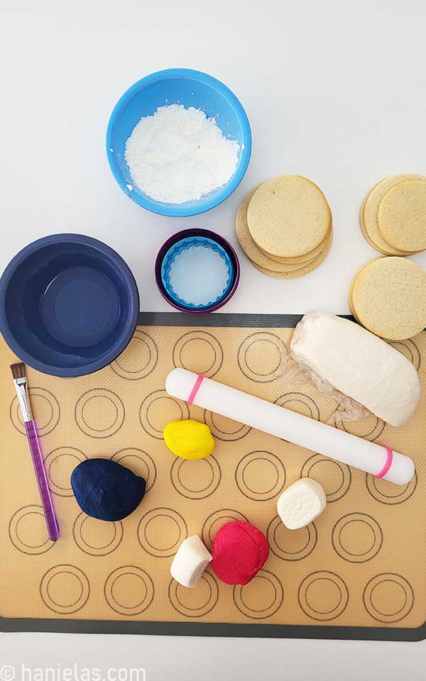 View from the above, undecorated cookies, a block of fondant, colored fondant pieces, a rolling pin, a silicone mat, and starch in a bowl.