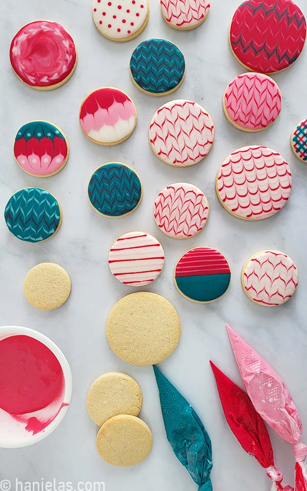 Small bowl with icing, undecorated round cookies, three piping bag filled with icing and lot of decorated cookies on a marble slab.