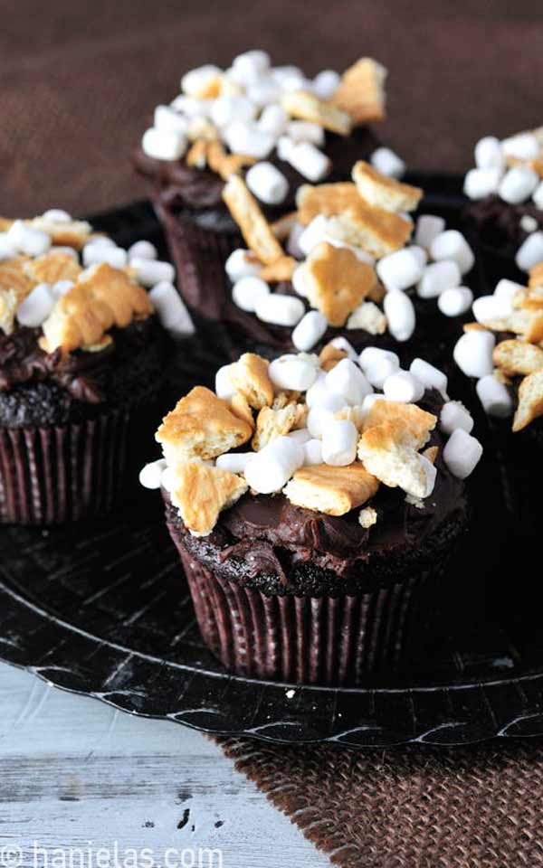 Cupcakes on black table, frosted with ganache and decorated with crushed cookies and marshmallows.