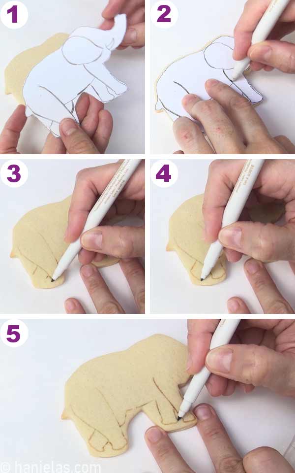 A cookie with a cardstock template on top of it, hand holding an edible marker, tracing outlines onto the cookie.