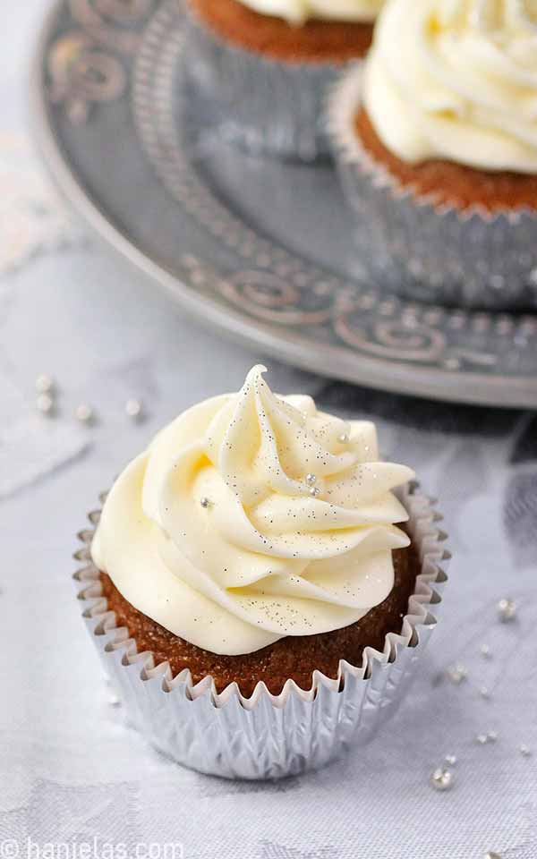 A golden colored cupcake in a silver cupcake liner with a frosting swirl dusted with silver nonpareils sprinkles.
