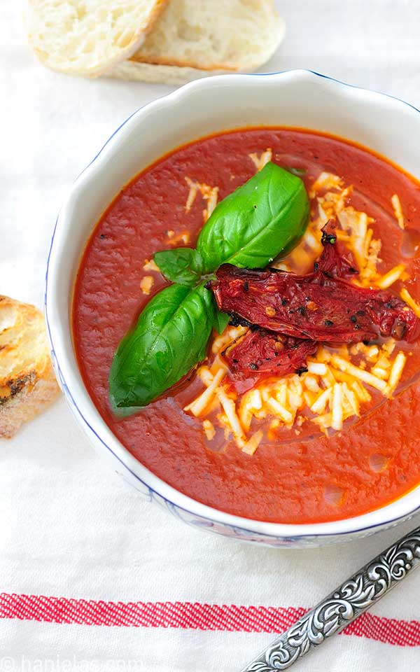 White bowl with tomato soup, topped with cheese and fresh basil leaves.