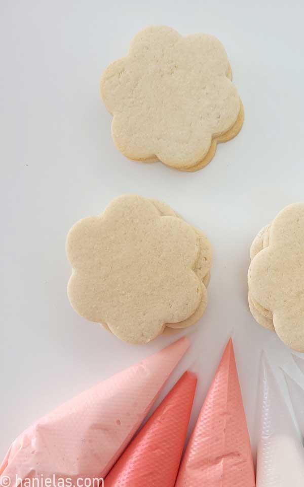 Undecorated flower cookies alongside piping bags filled with royal icing.