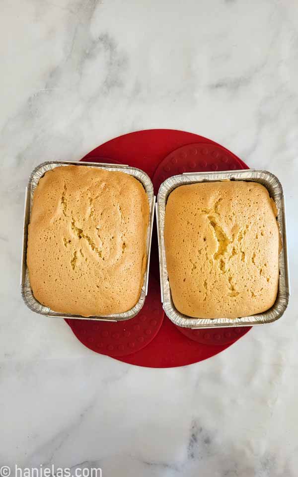 Two baked cakes in aluminum baking pans on a counter.