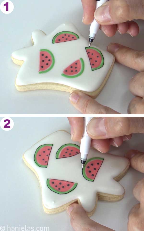 Hand holding a fine-tip edible marker, drawing outlines around wet-on-wet royal icing watermelons.