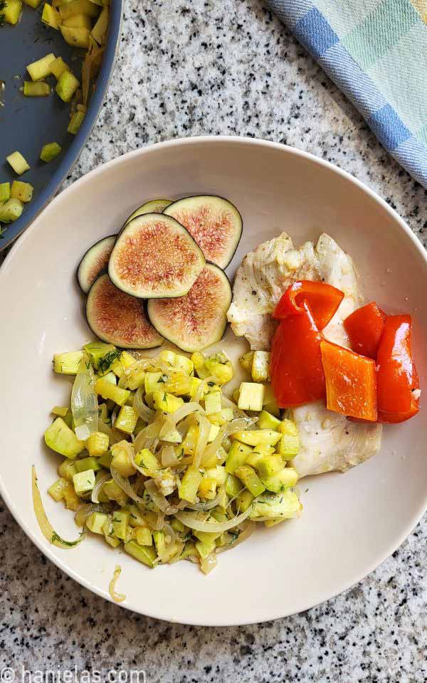 A bird's view of an ivory plate with sliced fig, cooked chicken and red pepper and diced cooked zucchini.