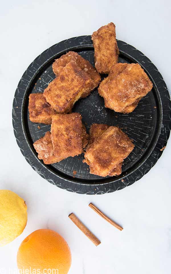 View from the above, black plate with fried cakes, beside a lemon, orange and cinnamon sticks.