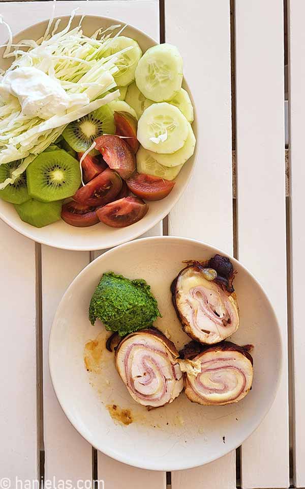 Two plates, one with sliced three chicken roll slices, on the side spinach pesto, and a a bowl with sliced cucumber, kiwifruit, tomatoes and shredded cabbage and a dollop of white greek yogurt.
